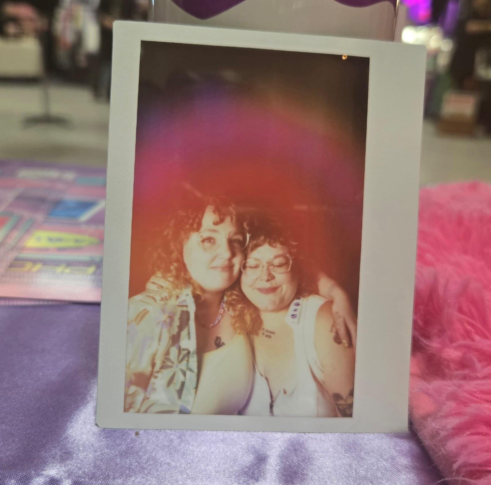 An aura portrait of two people cheek to cheek, wrapped in warm pink and gold light, propped up on a market table.